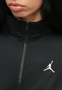 Tuvumā redzama melna jaka ar rāvējslēdzēju, uz krūtīm ir balts basketbola spēlētāja logo, kas lēkā, un daļēji redzams personas lūpu un zoda skats.