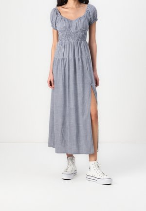 Frau trägt ein blau-weiß kariertes Midikleid mit Puffärmeln, seitlichem Schlitz und weißen High-Top-Sneakers vor schlichter Hintergrund.