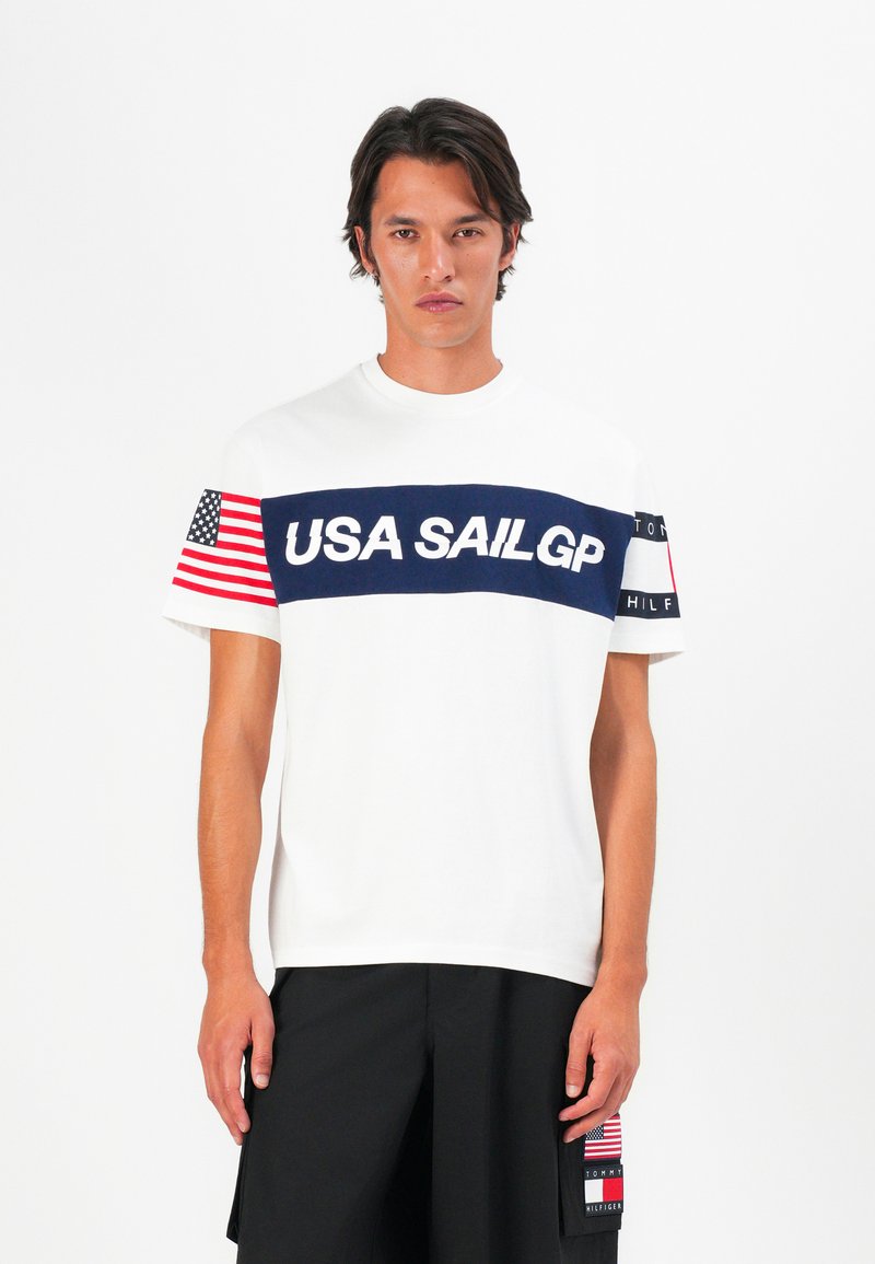 White t-shirt featuring a navy horizontal band with "USA SAILGP" text. Left sleeve has an American flag. Black shorts with a pocket patch.