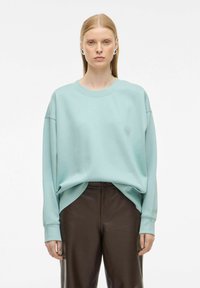 Donna che indossa una felpa oversized blu chiaro e pantaloni in pelle marrone scuro, in piedi su uno sfondo bianco semplice.