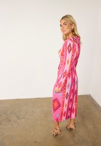 Γυναίκα με πολύχρωμο ροζ μακρύ φόρεμα γεωμετρικό μοτίβο και ψηλοτάκουνα σανδάλια με σχέδιο λεοπάρ, να στέκεται πλάγια σε τσιμεντένιο δάπεδο.