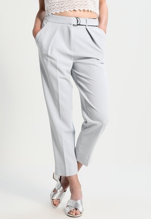 Frau trägt hellgraue, hoch taillierte, mit Gürtel versehene Hose mit Falten, in verkürzter Länge, und silberne Absatzsandalen, Hände in den Taschen.