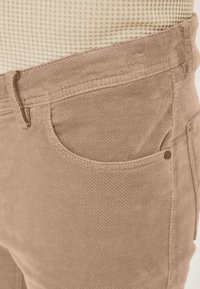 Beige Cord-Hose mit strukturierter Oberfläche, ausgestattet mit einer vorderen Tasche und einer kleinen Metallverzierung, dargestellt auf Taillenhöhe.