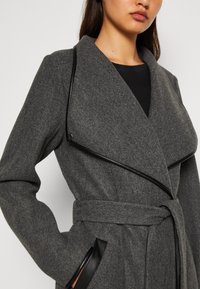 Harmaa päällystakki, valmistettu teksturoidusta kankaasta, jossa mustat nahkayksityiskohdat, syvä kaulus ja sidottu vyötärö. Mallissa on sivutaskut ja istuva leikkaus.