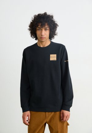 Black sweatshirt with a round neckline and long sleeves. It features a tan rectangular patch on the chest with branding. Textured fabric, casual fit.