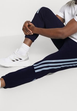 Man sitting on floor holding bent knee, wearing navy blue pants with light blue stripes, white socks, white Adidas sneakers, and a white shirt.