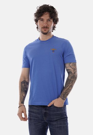 Uomo con capelli ricci e tatuaggi su entrambe le braccia che indossa una maglietta blu a maniche corte con un emblema e jeans scuri, in piedi contro uno sfondo bianco.