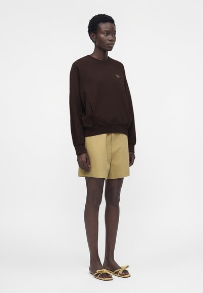 Γυναίκα που στέκεται φορώντας ένα σκούρο καφέ μακρυμάνικο φούτερ, μπεζ σορτς μέχρι το γόνατο και μπεζ σανδάλια με φιόγκους.