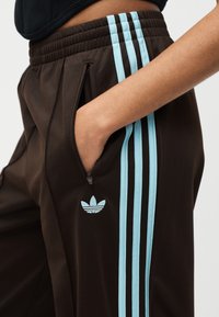 Άτομο που φοράει καφέ παντελόνια φόρμας Adidas με ανοιχτό μπλε ρίγες και λογότυπο, με το χέρι μέσα σε τσέπη με φερμουάρ στο πλάι.