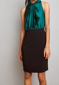 Tyrkysový saténový top s halter výstřihem a uvázaným výstřihem, spárovaný s přiléhavou černou sukní; hladká textura a čisté linie zdůrazňují siluetu.