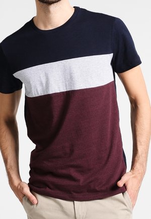 Uomo che indossa una t-shirt a maniche corte con scollo rotondo con blocchi di colore blu navy, bianco a righe e bordeaux, mani nelle tasche di pantaloni beige.