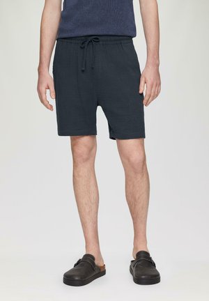 Shorts bleu marine texturés avec une taille élastique et un cordon de serrage ajustable. Portés avec des chaussures brunes à enfiler, debout sur un fond neutre.