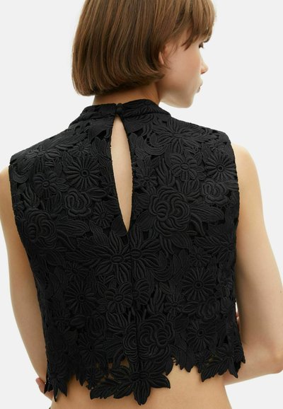 Frau trägt ein ärmelloses schwarzes Spitzenoberteil mit floralem Muster und Schlitzknopfverschluss hinten, cropped Länge, die Haut darunter zeigt.