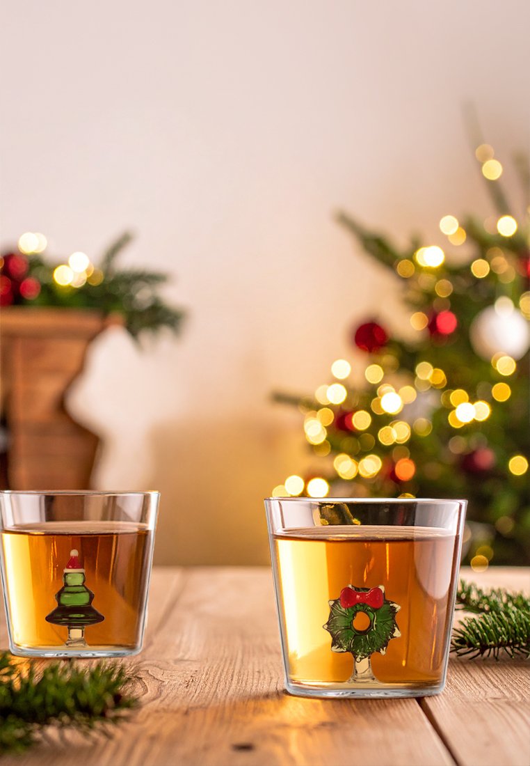 Zwei klare Gläser, gefüllt mit bernsteinfarbener Flüssigkeit, mit einem Weihnachtsbaum- und Kranzdesign am Fuß. Holztisch und festlicher Hintergrund.