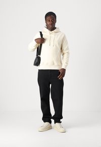 Felpa crema con testo in rilievo, borsa nera con tracolla, pantaloni neri e sneakers di colore chiaro. Design semplice e moderno.