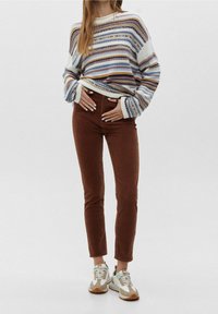 Sweter z dzianiny w paski w kolorach białym, niebieskim, fioletowym i żółtym, zestawiony z brązowymi spodniami z kordurowej tkaniny oraz jasnymi sneakersami z białymi i beżowymi akcentami.