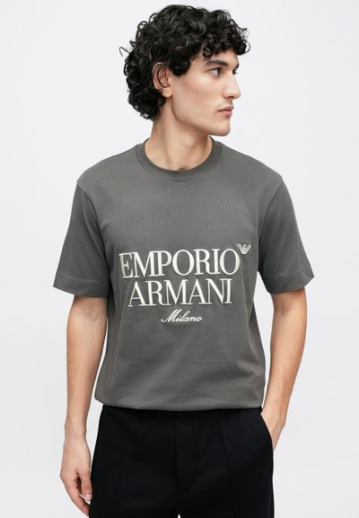 Sivá bavlnená tričko s bielym nápisom "EMPORIO ARMANI Milano", klasickým okrúhlym výstrihom a krátkymi rukávmi. Mäkký materiál a pohodlný strih.