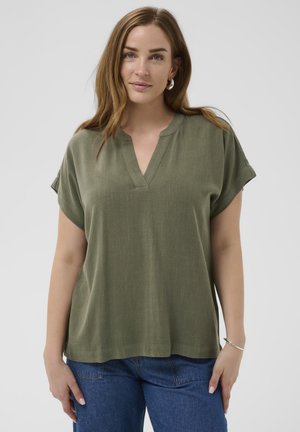 Femme aux longs cheveux châtain clair portant des boucles d'oreilles créoles argentées, un haut ample à manches courtes vert olive et un jean bleu, sur un fond uni.