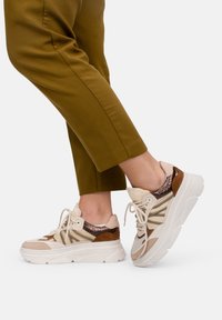 Beige en bruine chunky sneakers van mesh en suede, met textuuraccenten en witte rubberen zolen, gestyled met olijfgroene broeken.