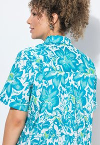 Camicia a maniche corte con un motivo floreale in teal e bianco. Presenta un colletto e una vestibilità rilassata con una texture leggera e liscia.