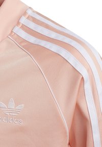Chaqueta rosa claro con diseño de tres rayas blancas y logotipo bordado, hecha de una tela suave con acentos texturizados a lo largo de las costuras.