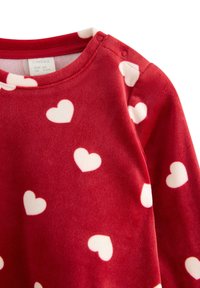 Red fleece garment with white heart patterns, rounded neckline, and snap buttons on the shoulder for easy dressing. Soft texture.