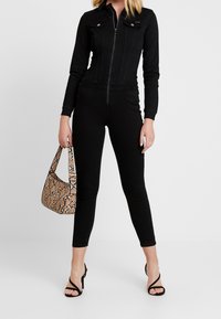 Black denim jumpsuit with zip closure, long sleeves, and two chest pockets; paired with a tan snakeskin handbag and black strappy heels.