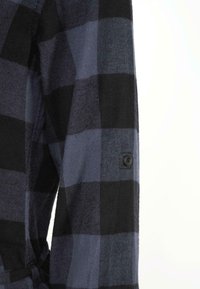 Dettaglio di una camicia a quadri nera e blu navy con polsino abbottonato, texture tessuta e un motivo a quadri prominente su un tessuto morbido.