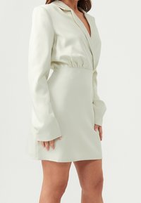 Femme portant une mini-robe crème à manches longues avec un col en V profond et une taille froncée, debout devant un fond blanc uni.