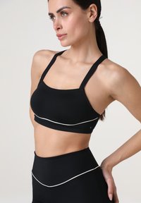 Reggiseno sportivo nero con spalline spesse e una texture liscia. Linea accentuata bianca lungo il fondo. Design aderente, adatto per abbigliamento sportivo.