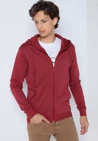 Haut de hoodie zippé rouge avec capuche à cordon, texture lisse et un logo discret sur la poitrine. Associé à un t-shirt blanc et un pantalon beige.