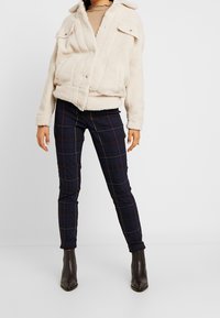 Veste en polaire crème avec fermeture éclair à l'avant et deux poches poitrine, associée à un pantalon ajusté à carreaux navy et des bottines à talons.