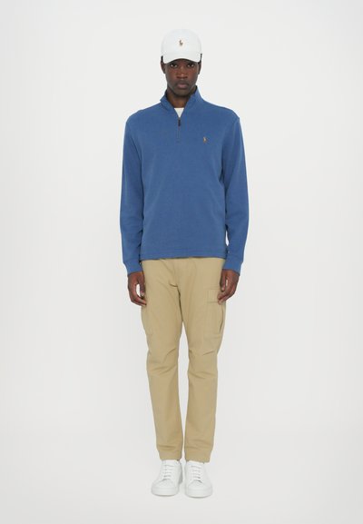 Blauwe trui met een kwart-rits kraag, beige cargobroek en witte sneakers, tentoongesteld tegen een effen achtergrond.