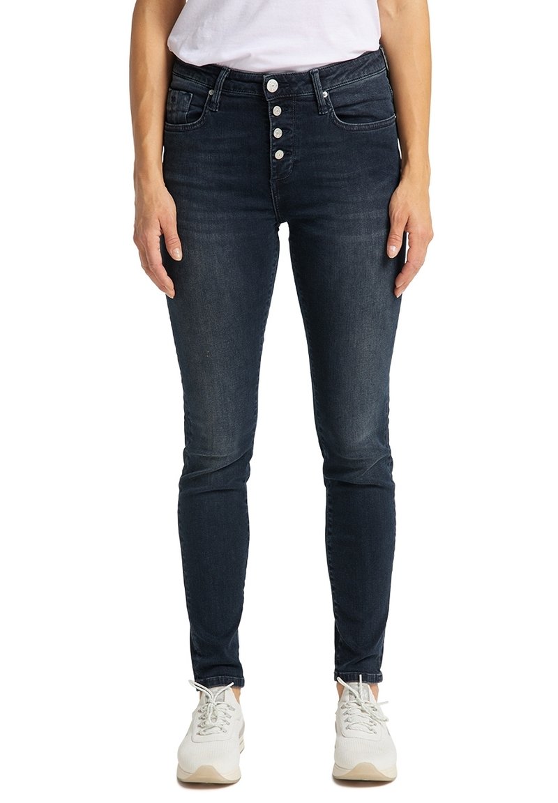 Femme portant un jean skinny foncé avec quatre boutons métalliques visibles et des baskets blanches, debout devant un fond blanc.