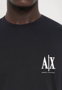 Marineblå bomulls t-skjorte med rund hals og en hvit brodert logo, "A|X," på venstre bryst. Glatt tekstur.