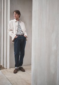 Jeune homme debout dans un couloir aux tons neutres, portant une veste claire, une chemise rayée, un jean foncé et des chaussures marron, les mains dans les poches.