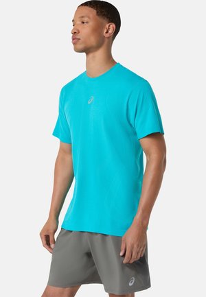 Mann trägt ein hellblaues kurzärmeliges Sportshirt und graue Shorts, steht in Dreiviertelansicht vor weißem Hintergrund.