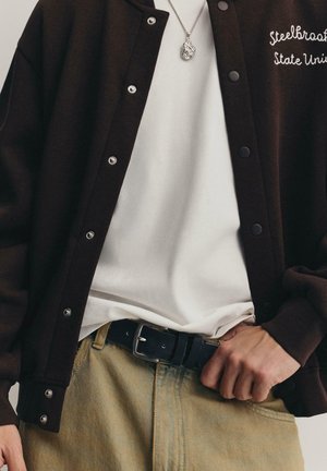 Braune Collegejacke mit Druckknöpfen, weißes T-Shirt darunter, schwarzer Gürtel und hellbraune Hose. Beinhaltet eine silberne Anhänger-Halskette.