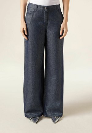 Jean en denim foncé à jambes larges porté avec des chaussures argentées à bouts pointus, associé à un haut blanc rentré, sur une personne immobile.