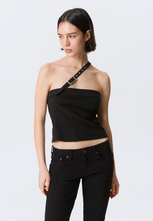 Kobieta o krótkich ciemnych włosach, ubrana w czarny, bezrękawnikowy top z paskiem z klamrą na ramieniu oraz czarne spodnie, stojąca na jednolitym tle.