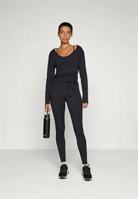 Schwarzes langärmliges Wickeloberteil mit einem Bund am Taillenbereich, kombiniert mit schwarzen Leggings und schwarzen Turnschuhen, hält eine schwarze Wasserflasche.