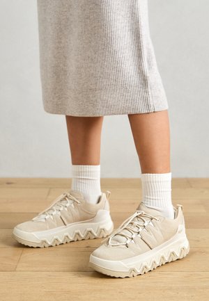 Beige sportssko med en struktureret, glat overdel, der har elastiske snørebånd og en takket sål. Båret med hvide ribbede sokker.