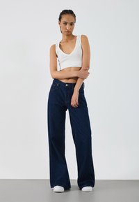 Top scurt alb, blugi evazați de culoare bleumarin și adidași albi. Material neted, design clasic din denim cu talie înaltă și buzunare.