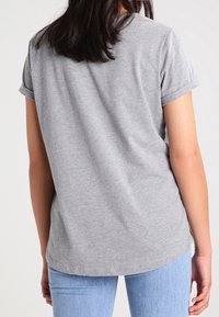 Camiseta de algodón gris con mangas cortas enrolladas y un dobladillo redondeado. Tiene una textura suave y se ajusta holgadamente en la parte de atrás. Se lleva con jeans azules.