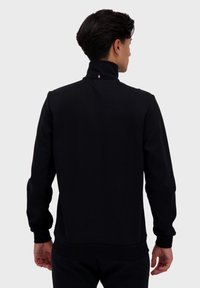 Felpa nera con colletto alto, polsini e orlo a costine. Presenta una piccola etichetta tricolore sulla parte posteriore del collo. Texture liscia e morbida.