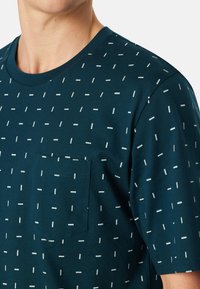 Camiseta de color verde azulado oscuro hecha de algodón con cuello redondo, que presenta un bolsillo y patrones rectangulares blancos dispersos. Textura suave.