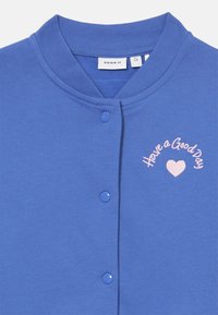 Blå bomullskardigan med rund hals, knappelukking og brodert tekst "Ha en god dag" med en rosa hjerte detalj.