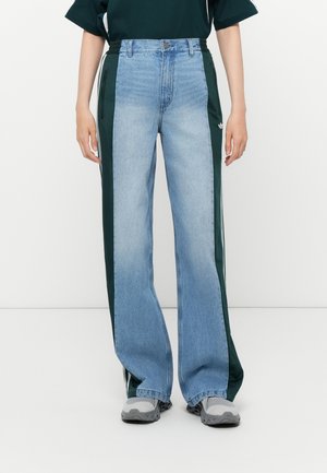Jean large en denim bleu avec des panneaux latéraux verts, dotés d'une taille haute et de poches zippées, associés à des baskets grises.