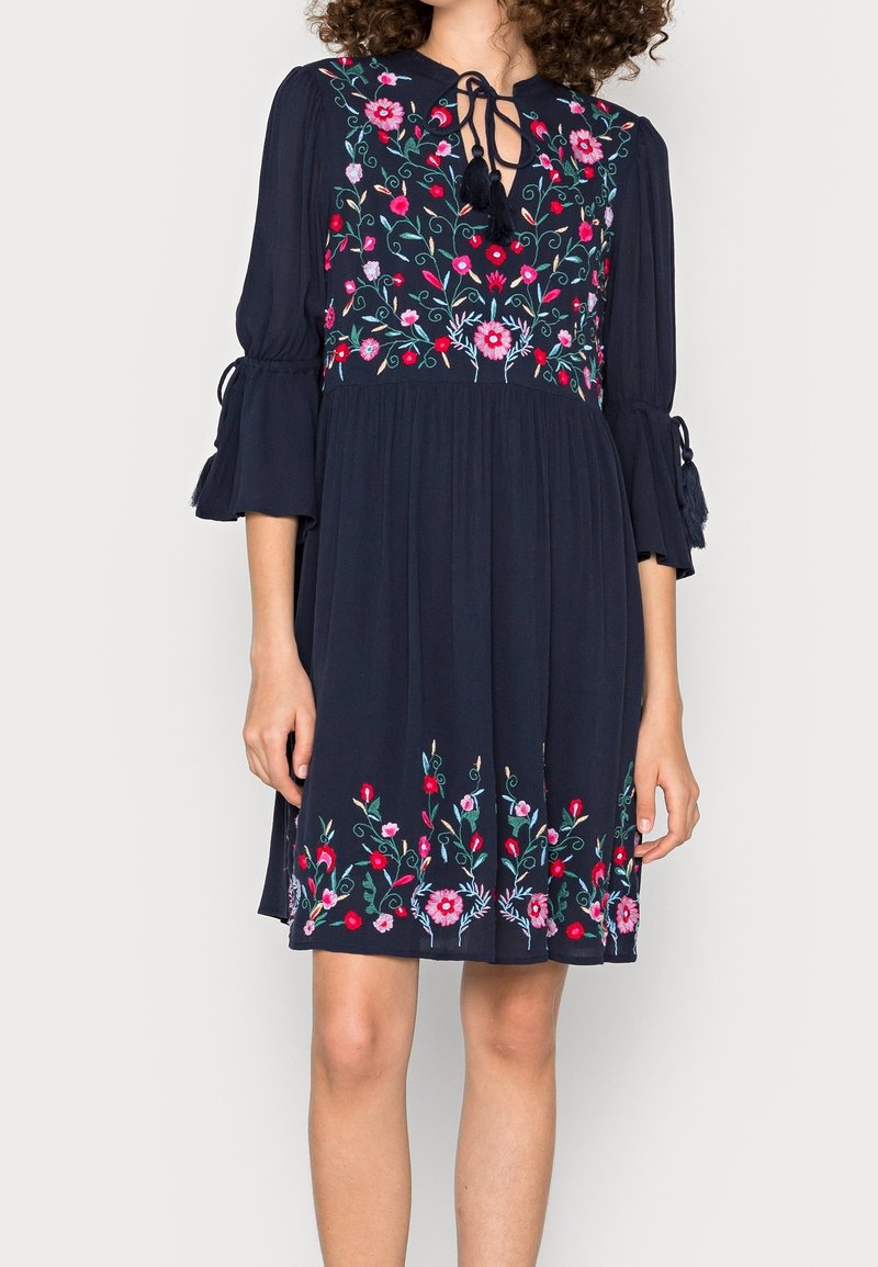 Mörkblå knälång klänning med röd och rosa blommig broderi, trekvarts ärmar med klockform och flätade fransar, samt en knytning vid nyckelhålet.