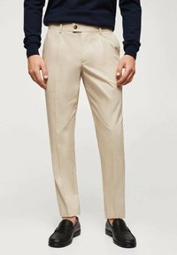 Pantaloni beige con vestibilità sartoriale, dotati di design a gamba dritta senza pieghe, tasche frontali e chiusura con un solo bottone. Indossati con mocassini neri.